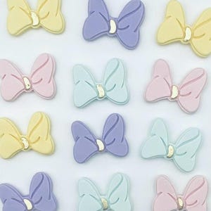 May include: A collection of pastel-colored bow-shaped embellishments. The bows are in shades of yellow, lavender, pink, and light blue, each with a small gold-colored center. These decorative bows are arranged in a grid pattern.
