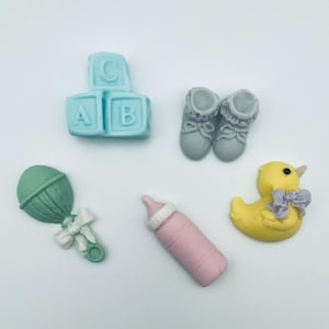 May include: An assortment of baby-themed decorations. Includes light blue alphabet blocks, gray baby booties, a green rattle, a pink baby bottle, and a yellow duck with a purple bow.