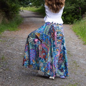 May include: A colourful, patterned maxi skirt in shades of blue, purple, and pink, worn with a white top. The person is standing on a gravel path surrounded by trees. The skirt's pattern is intricate.