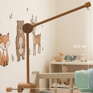 Op de afbeelding: Een houten baby mobile arm, 45,5 cm lang, bevestigd aan een wieg. De arm heeft een haak om speelgoed op te hangen. De wieg klem is 32,5 cm breed. De achtergrond toont muurstickers met bosdieren.