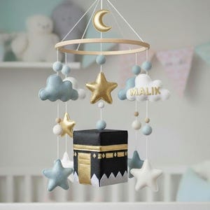 May include: A baby mobile featuring a wooden hoop with hanging felt decorations. Includes gold stars, a crescent moon, blue clouds, and a black cube with gold accents. The name "Malik" is embroidered on a cloud.
