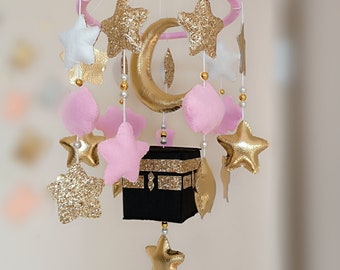 May include: A pink and gold baby mobile with stars, a crescent moon, and a small black cube with gold trim. The mobile is hanging from a pink ring.