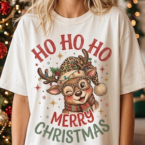 Peut inclure: Un t-shirt blanc avec une illustration de renne portant un bonnet à motif léopard, des lunettes et une écharpe. Le texte HO HO HO est inscrit au-dessus et MERRY CHRISTMAS en dessous en lettres rouges et vertes avec des étoiles.