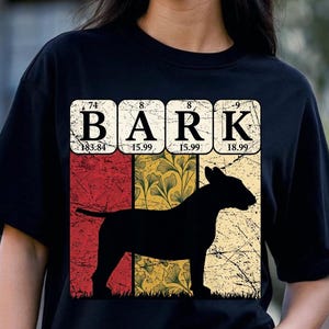 Peut inclure: Un t-shirt noir avec un motif graphique représentant le mot BARK stylisé comme des éléments du tableau périodique. Sous le texte se trouve la silhouette noire d'un chien sur un fond de bandes verticales rouges et jaunes à motifs de feuilles.