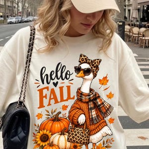 Peut inclure: Un sweat-shirt à col rond couleur crème avec un motif représentant une oie portant un nœud à imprimé léopard, des lunettes de soleil, une écharpe à carreaux et un pull orange, tenant un sac à main léopard. Le texte hello FALL est inscrit.