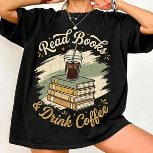 Peut inclure: Un t-shirt noir à manches courtes avec une illustration d'une pile de quatre livres surmontée d'un café glacé dans un gobelet en plastique. Le texte "Read Books & Drink Coffee" est imprimé en arc de cercle dans une police crème.