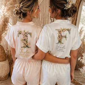 May include: Two women in matching satin pyjama sets, one pale pink, one white. Each set features a floral initial design on the back, with a name and date. The women are embracing, their backs to the camera.