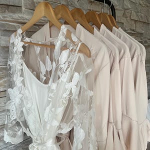 May include: A row of five light beige robes hanging on wooden hangers. The first robe features a sheer white lace overlay with floral embroidery and a matching tie belt. The remaining four robes are made of a solid, smooth, light beige fabric.