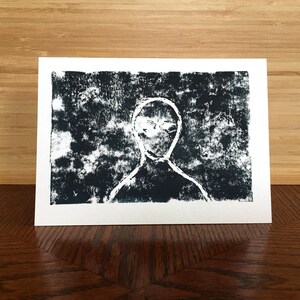 May include: A black and white print on white cardstock. The print features a stylized figure with a white outline against a textured black background. The figure has a simple, minimalist design, with a visible head and shoulders.