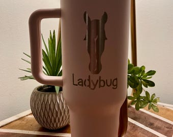 May include: A matte light pink insulated tumbler with a handle and a lid. The front features a laser-engraved silhouette of a horse head positioned above the text "Ladybug" in a matching font. A light pink straw protrudes from the top lid.