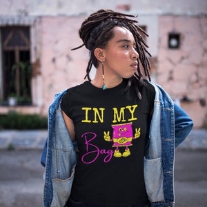 May include: Black t-shirt with the text "IN MY Bag" in yellow and pink. A cartoon graphic of a bag with a face and peace signs is below the text. The shirt is worn with a denim jacket and dark blue jeans.