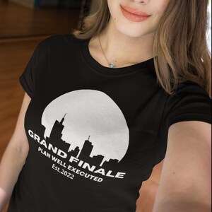 Może przedstawiać: Czarny t-shirt z białym nadrukiem przedstawiającym panoramę miasta w okręgu. Pod spodem znajduje się napis "GRAND FINALE PLAN WELL EXECUTED Est.2022". Koszulka ma okrągły dekolt.