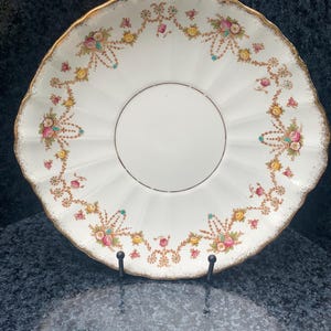 May include: An antique white plate with a scalloped edge and gold trim. The plate has a floral design in pink, yellow, and teal. The plate is displayed on a black metal stand.