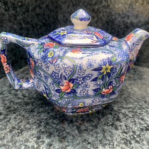 May include: A ceramic teapot featuring a vibrant blue base colour with an intricate pattern of small white flowers, green vines, and larger pink and yellow floral accents. The teapot includes a matching lid with a small knob handle and a spout.