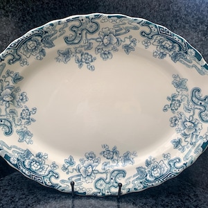 May include: An oval, white ceramic serving platter with a teal floral design around the rim. The platter has a scalloped edge and is supported by a black metal stand. The floral pattern includes stylized flowers and decorative elements.