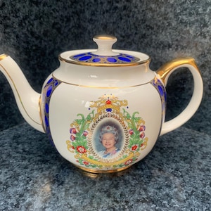 Puede incluir: Una tetera de porcelana blanca con un retrato conmemorativo de la reina Isabel II rodeado por una corona floral y el texto 1953-2003. La tetera está decorada con detalles en azul y oro en la tapa, el asa, el pico y el cuerpo.
