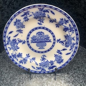 Könnte beinhalten: Ein runder Keramikteller mit cremefarbener Basis und blauem Transferdruckmuster. Die Mitte zeigt einen Blumenkorb innerhalb einer kreisförmigen Umrandung, während der Rand mit rankenden Blumenmotiven und einem geometrischen Rautenmuster verziert ist.