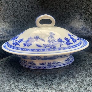 May include: A white porcelain serving dish with a lid, featuring a blue floral pattern and a geometric border. The oval-shaped dish has a pedestal base and a loop handle on the lid, decorated with blue botanical motifs and intricate trim.