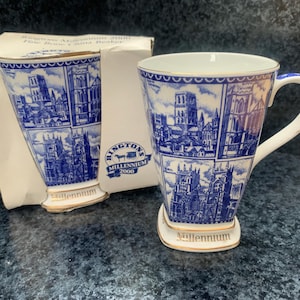 May include: A white fine bone china beaker with a blue and white transfer print featuring architectural illustrations of cathedrals. The beaker has a gold rim and base, with the word Millennium printed in gold. It is accompanied by its original box.