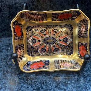 Peut inclure: Un plat rectangulaire en porcelaine aux bords festonnés, présentant une palette de couleurs bleu profond et or. L'intérieur est orné de motifs floraux complexes en orange, blanc et or, accentués par des motifs géométriques sur le bord.