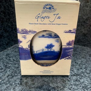 May include: A rectangular cardboard box containing a porcelain ginger jar. The box is cream-colored with blue landscape illustrations and text reading Ringtons Ginger Jar, Finest Dark Chocolates with Stem Ginger Centres, and Porcelain Produced Exclusively for Ringtons.