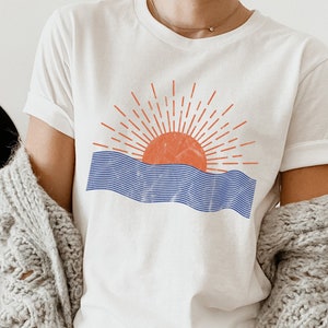 May include: White t-shirt with a graphic of a red sun setting over blue waves. The sun has a textured look and the waves are made of thin blue lines.