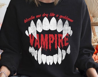 Könnte beinhalten: Ein schwarzes Sweatshirt mit Rundhalsausschnitt und einer Grafik aus weißen Vampirzähnen sowie dem roten Schriftzug "bleedin me dry like a goddamn VAMPIRE" auf der Brust. Das Design zeigt eine abgenutzte Textur auf den weißen Zähnen und tropfende rote Buchstaben.