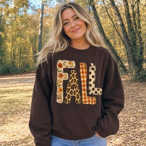Puede incluir: Una sudadera de cuello redondo en color marrón oscuro con la palabra FALL impresa en letras grandes. Cada letra tiene un patrón distinto: la F tiene un estampado floral, la A un estampado de leopardo, la L un cuadro vichy naranja y la L puntos marrones.