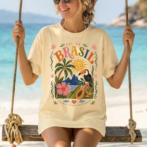 Puede incluir: Camiseta color crema con un diseño gráfico colorido que presenta la palabra "BRASIL" en naranja y verde, un tucán, palmeras y flores tropicales. La camiseta tiene mangas cortas y un corte holgado.