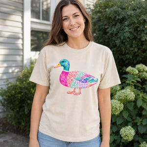 Pode incluir: Uma t-shirt bege de manga curta com um gráfico colorido de um pato-real. A ilustração do pato apresenta uma mistura vibrante de padrões em rosa, azul, verde e roxo, incluindo bolinhas e xadrez, com um bico amarelo brilhante.