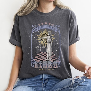Könnte beinhalten: Dunkelgraues T-Shirt mit einem Waage-Sternzeichen-Design. Die Grafik zeigt eine Frau mit Waage, eine Sonne und den Text "LIBRA SEP 23 - OCT 22" im Vintage-Stil. Das Shirt hat hochgekrempelte Ärmel und eine lockere Passform.