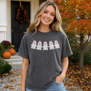 May include: A charcoal gray short-sleeve t-shirt featuring a graphic print of five white ghosts holding hands in a row. Each ghost has a small pink bow on its head. The shirt has a crew neckline and a relaxed fit, made of a washed-out fabric.