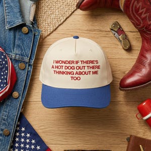 May include: A baseball cap with a cream-colored crown and a blue brim. The cap features the text "I WONDER IF THERE'S A HOT DOG OUT THERE THINKING ABOUT ME TOO" in red. The cap is on a wooden surface.