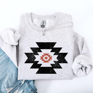 Könnte beinhalten: Ein graues Sweatshirt mit einem geometrischen Muster in Schwarz und Rot. Das Muster ist ein stilisiertes Muster aus Quadraten und Dreiecken.