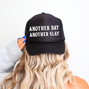 Op de afbeelding: Zwarte truckerpet met witte tekst "ANOTHER DAY ANOTHER SLAY". De pet heeft een mesh achterkant en een gebogen klep. De afbeelding toont de achterkant van het hoofd van een persoon met blond haar en een hand met blauwe nagellak.
