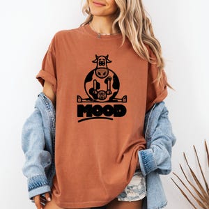 Könnte beinhalten: Rostfarbenes T-Shirt mit einem schwarzen Aufdruck einer sitzenden Kuh und dem Wort "MOOD" in fetter Schrift. Das Design der Kuh zeigt sie mit gespreizten Beinen. Das T-Shirt hat kurze Ärmel und eine lockere Passform.