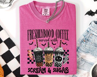 May include: A pink t-shirt with the text "FRESHLYBOO'D COFFEE served with SCREAM & SUGAR." The shirt features three coffee cup designs with Halloween-themed graphics. A silver chain is draped over the shirt.