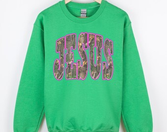 Puede incluir: Una sudadera verde brillante con la palabra "JESUS" en letras grandes y en bloque en la parte delantera. Las letras están rellenas con un patrón de camuflaje y delineadas en rosa vibrante. La sudadera está colgada en una percha de madera.