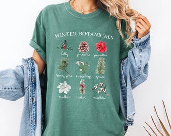 May include: A forest green short-sleeve t-shirt featuring the text "WINTER BOTANICALS" above a grid of nine illustrations of plants including holly, pinecone, poinsettia, torrey pine, snowdrop, spruce, mullein, cedar, and mistletoe with labels.