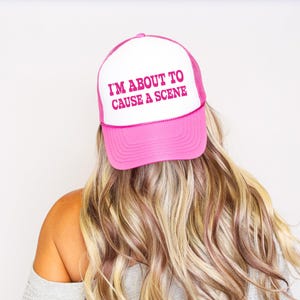 Może przedstawiać: Różowo-biała czapka typu trucker z różowym napisem "I'M ABOUT TO CAUSE A SCENE". Czapkę nosi osoba z blond i brązowymi włosami. Czapka ma różowy daszek i siatkową tylną część.