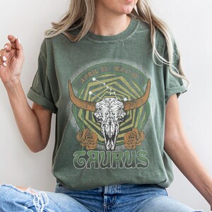 May include: Green t-shirt featuring a Taurus zodiac design. The graphic includes a bull skull with horns, the word "TAURUS", and the dates "APRIL 20 - MAY 20". The design is surrounded by floral and geometric elements.