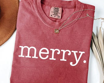 Puede incluir: Una camiseta de manga corta color rojo ladrillo con cuello redondo y la palabra "merry." impresa en letras blancas sin remates en el pecho. La camiseta tiene una etiqueta de la marca Comfort Colors en el cuello con la talla S-P-CH visible.
