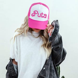 May include: A pink and white trucker hat with the word "Puta" in pink script. The hat has a mesh back and a curved brim. The hat is being held up by a person.