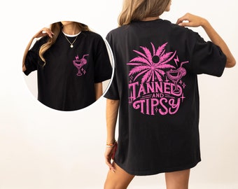 May include: Black oversized t-shirt with a pink palm tree and cocktail graphic on the back. The text "Tanned and Tipsy" is written below the graphic. A pink cocktail graphic is on the front of the shirt.