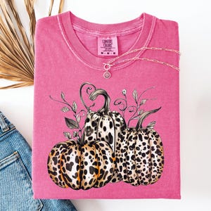 May include: A pink Comfort Colors t-shirt featuring a graphic of three pumpkins with leopard print patterns. A silver necklace with a sunburst pendant is draped across the shirt. The shirt is folded, with a pair of blue jeans visible.