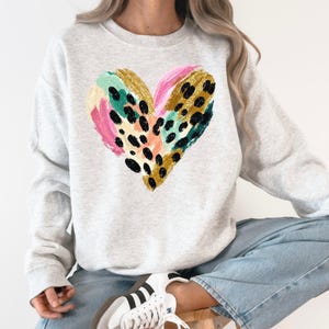 Puede incluir: Sudadera gris claro con un diseño de corazón colorido. El corazón está compuesto por pinceladas en rosa, dorado, turquesa y estampado de leopardo negro. La sudadera es de estilo casual y cómodo.