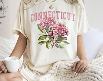 May include: Cream-coloured t-shirt with the word "CONNECTICUT" in pink block letters above a watercolour illustration of pink flowers and green leaves. The shirt has short sleeves and a relaxed fit.