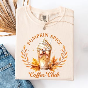 May include: Cream-colored t-shirt featuring a watercolor illustration of a pumpkin spice latte with whipped cream, a bow, and fall leaves. The text "Pumpkin Spice Coffee Club" is printed around the image. A silver necklace is draped across the shirt.
