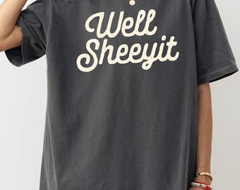 May include: Dark gray t-shirt with the text "Well Sheeeyit" in a cream-colored, cursive font. The shirt has short sleeves and a relaxed fit. The model is wearing a white undershirt and gold jewelry.