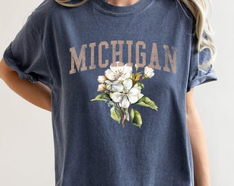 May include: Navy blue t-shirt with the word "MICHIGAN" in a distressed, tan font. Below the text is a watercolor illustration of white flowers with green leaves. The shirt has short sleeves.
